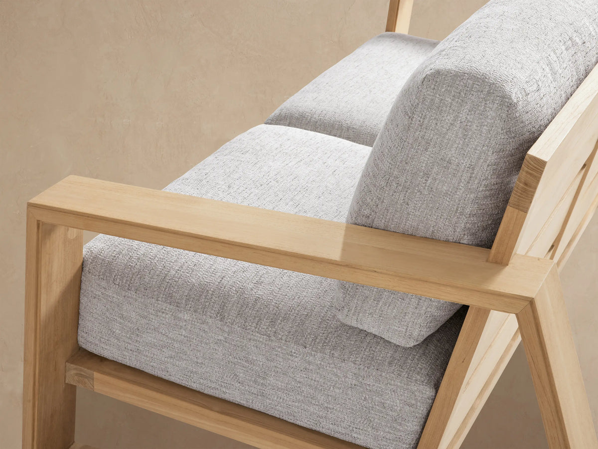 Canyon Outdoor Teak Sofa, 2-seater with light teak frame, angled open arms, and light grey cushions on textured stone.