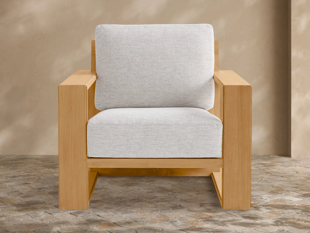 Canyon Outdoor Teak Lounge Chair with a light wood frame and thick light grey cushions, angled on a stone patio against a beige wall.