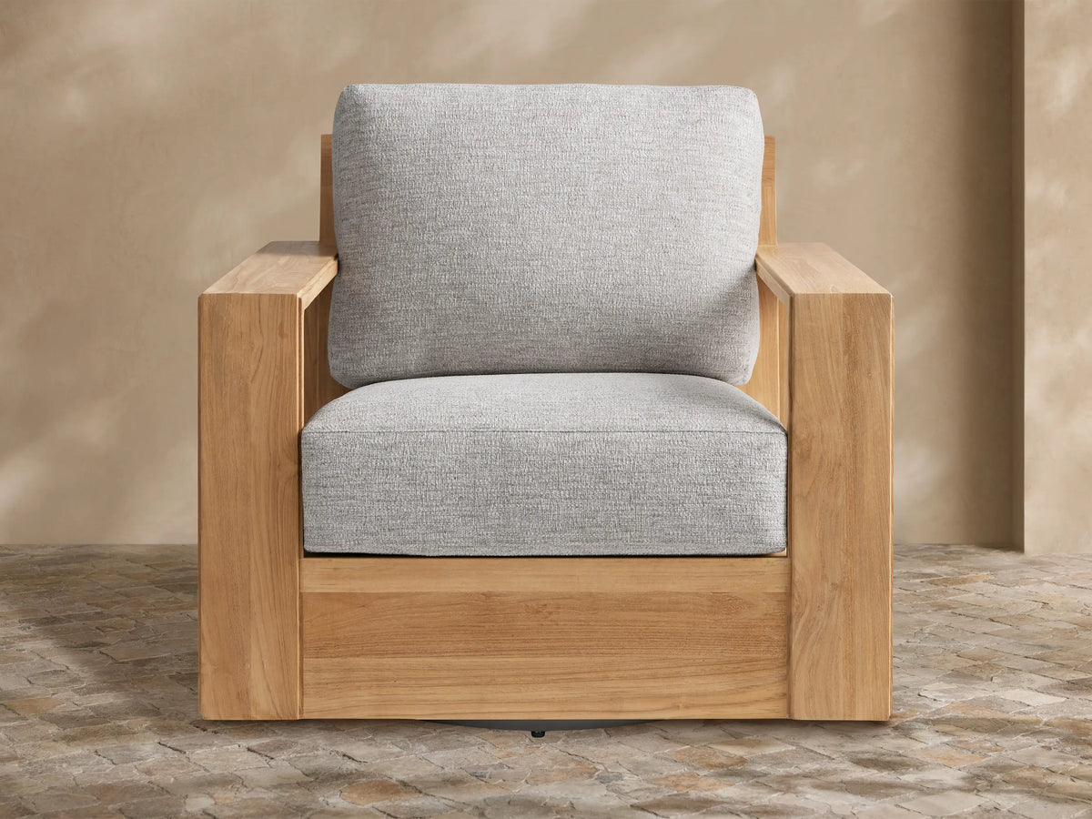 Canyon Outdoor Teak Swivel Chair with a modern, natural teak wood frame and light gray cushions, shown on a stone patio.