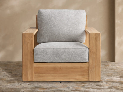 Canyon Outdoor Teak Swivel Chair with a modern, natural teak wood frame and light gray cushions, shown on a stone patio.
