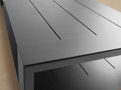 Canyon Outdoor Aluminum Coffee Table, dark grey, with a slatted top and lower shelf, on a stone patio against a tan wall.