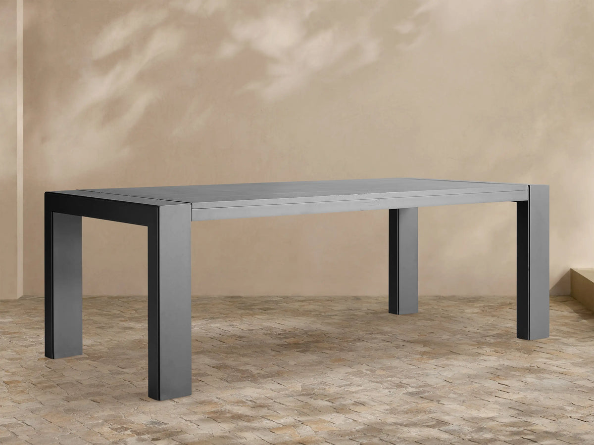 Dark gray rectangular Canyon Outdoor Aluminum Dining Table with slatted top and thick legs, on a light-colored paved patio.