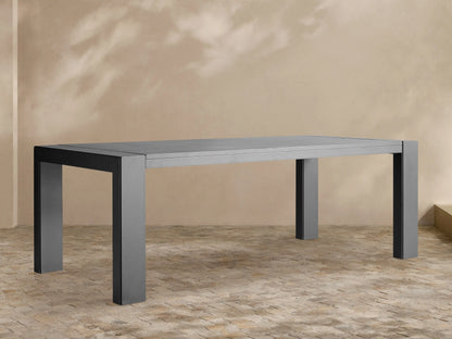 Dark gray rectangular Canyon Outdoor Aluminum Dining Table with slatted top and thick legs, on a light-colored paved patio.