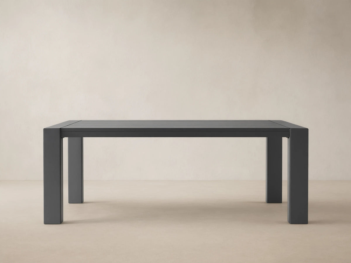 Dark gray rectangular Canyon Outdoor Aluminum Dining Table with slatted top and thick legs, on a light-colored paved patio.