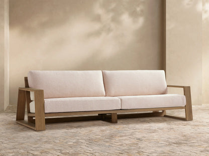 Front view of Canyon 2-seater Outdoor Teak Sofa, natural teak frame, light cream cushions, on a patio with a beige wall.