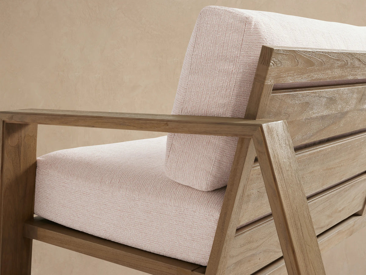 Canyon Outdoor Teak Sofa, 2-seater, natural teak frame and light beige cushions, set on a stone patio against a beige wall.