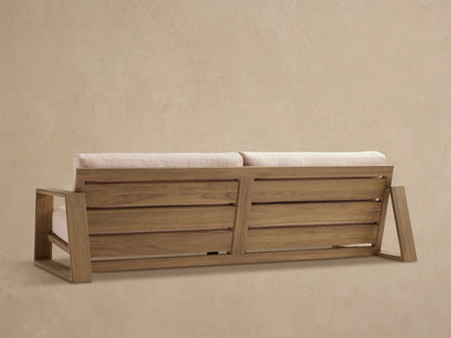 Canyon Outdoor Teak Sofa, angled close-up. Light gray-brown teak frame with textured grain, light pink woven cushions.