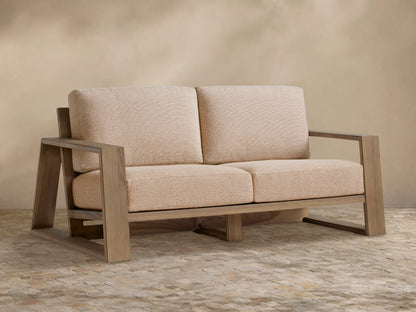 Canyon Outdoor Teak Sofa, a 2-seater with light teak wood frame and beige textured cushions, on tiled floor against light wall.