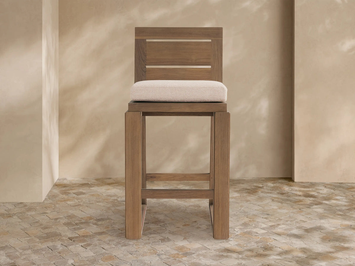 Canyon Outdoor Teak Barstool, light brown teak wood frame with slatted back and beige cushion, on tiled patio.