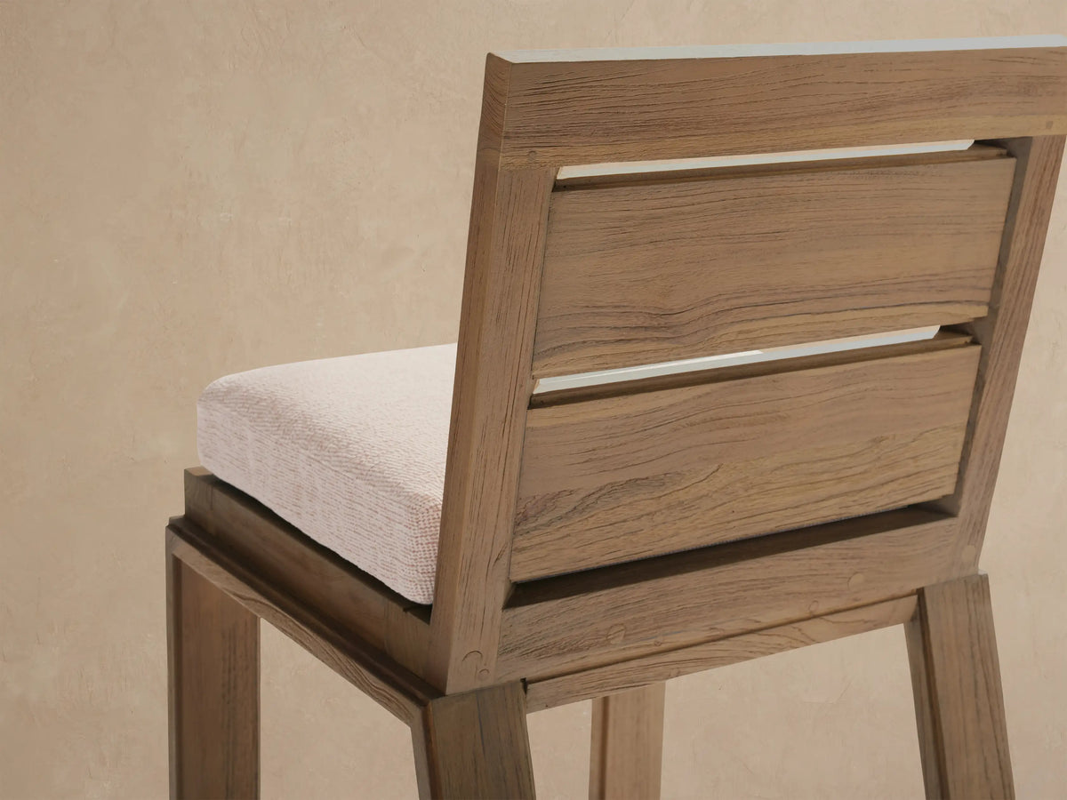 Front view of a natural teak Canyon Outdoor Barstool with a light beige cushion, against a textured wall on a tiled patio.
