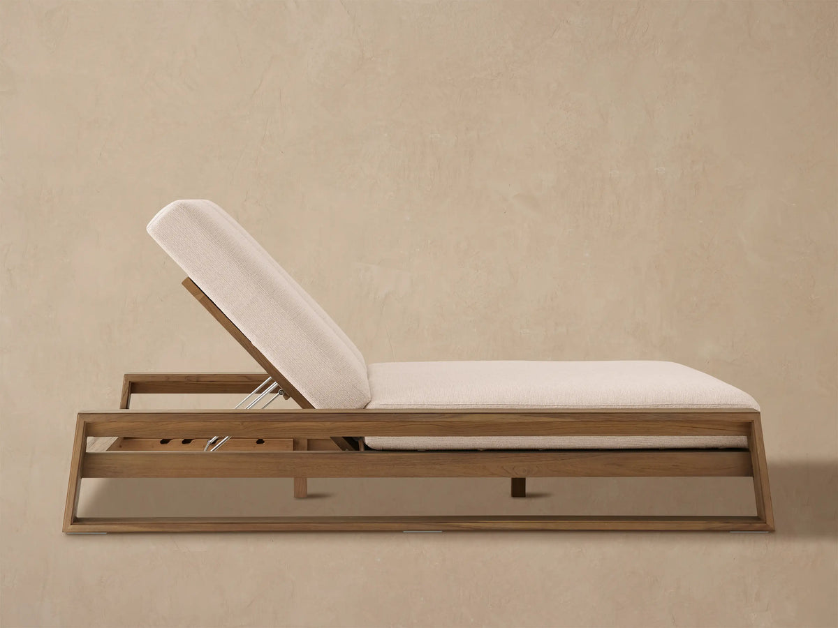 Canyon Outdoor Teak Double Chaise with light beige cushions on a stone patio against a tan wall, left backrest raised.