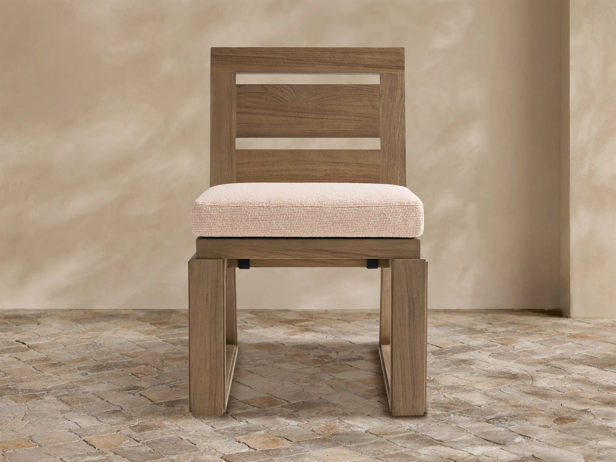 Canyon Outdoor Teak Dining Side Chair in natural finish with slatted back and light beige cushion, on a textured stone floor.