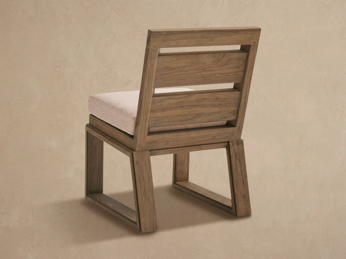 Canyon Outdoor Teak Dining Side Chair, angled view. Rustic brown teak frame with two open slats on back, light beige cushion.