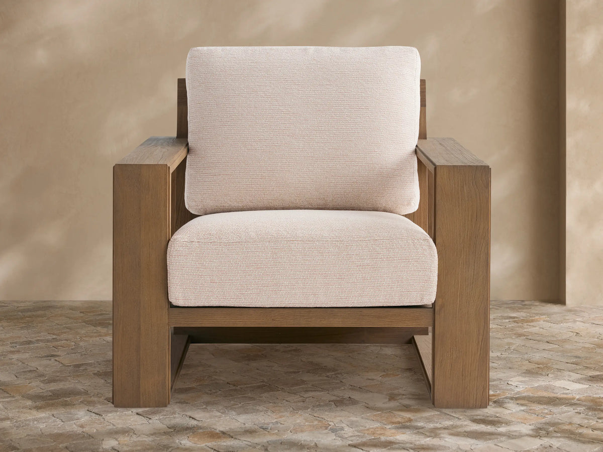 Canyon Outdoor Teak Lounge Chair. Modern, light brown teak frame, thick textured off-white cushions, on a patterned stone patio.