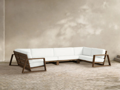 Canyon Outdoor Teak 4-piece Sectional, L-shaped with open-frame teak arms, white cushions on a stone patio by a light wall.