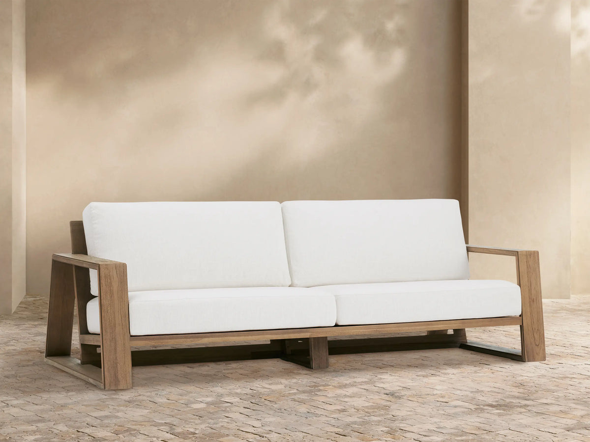 Canyon Outdoor Teak Sofa, front view, with light brown teak frame and two white seat/back cushions on a stone patio.