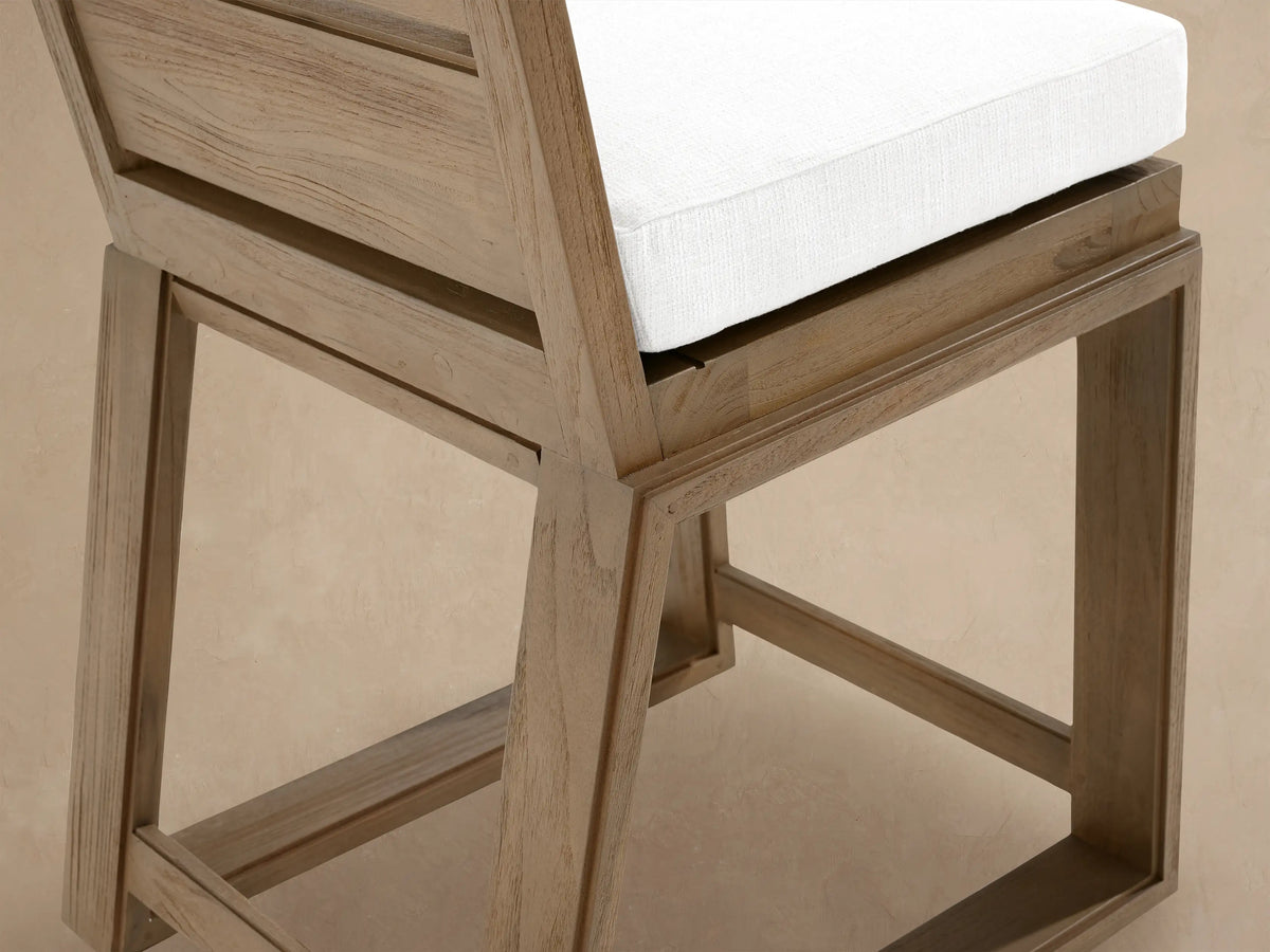 Canyon Outdoor Teak Barstool, light brown wood, white cushion, slatted backrest, front view, against a textured wall.