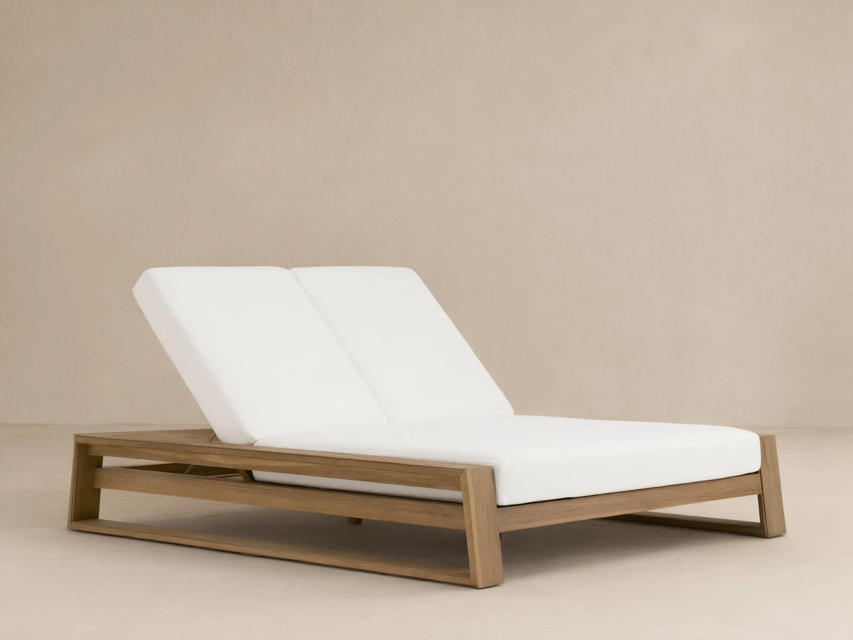 Canyon Outdoor Teak Double Chaise with light brown wood frame & white cushions. Backrests slightly raised, on a patio.