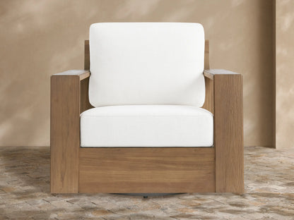 Canyon Outdoor Teak Swivel Chair with a modern, blocky natural teak frame & thick white cushions, angled on a tiled patio.