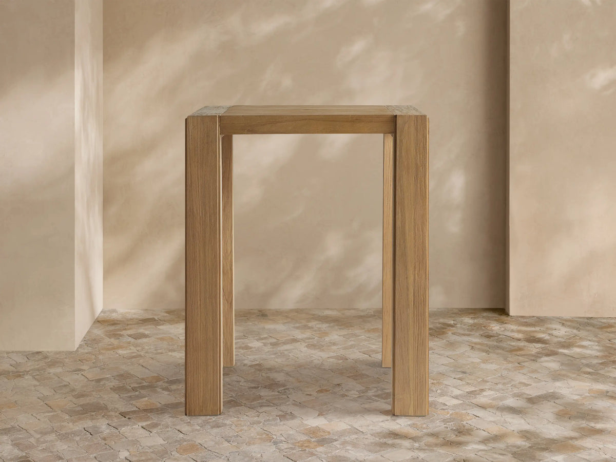 Light brown, rectangular Canyon Outdoor Teak Bar Table with natural wood grain and sturdy square legs, standing against a neutral wall.