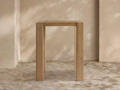 Light brown, rectangular Canyon Outdoor Teak Bar Table with natural wood grain and sturdy square legs, standing against a neutral wall.