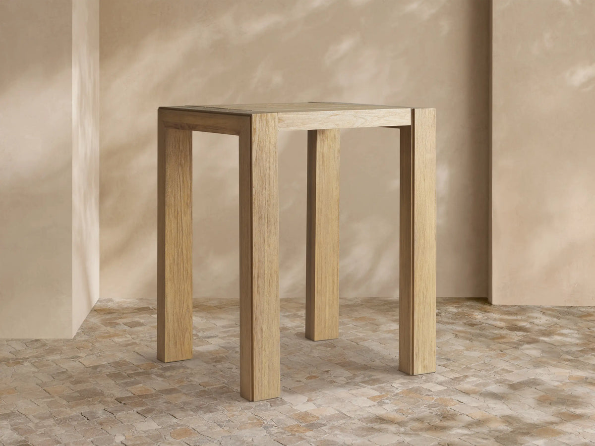Light brown, rectangular Canyon Outdoor Teak Bar Table with natural wood grain and sturdy square legs, standing against a neutral wall.