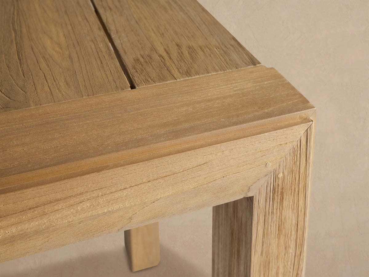 Square Canyon Outdoor Teak Bar Table with a slatted top & four sturdy square legs, light natural wood finish, on a rustic tiled floor.