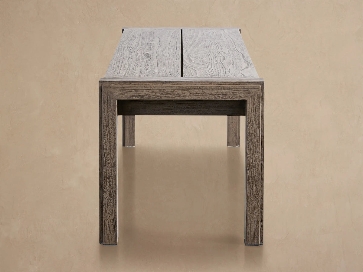 Rectangular Canyon Outdoor Teak Dining Bench in distressed gray-brown, with sturdy block legs, on a tiled patio against a light wall.