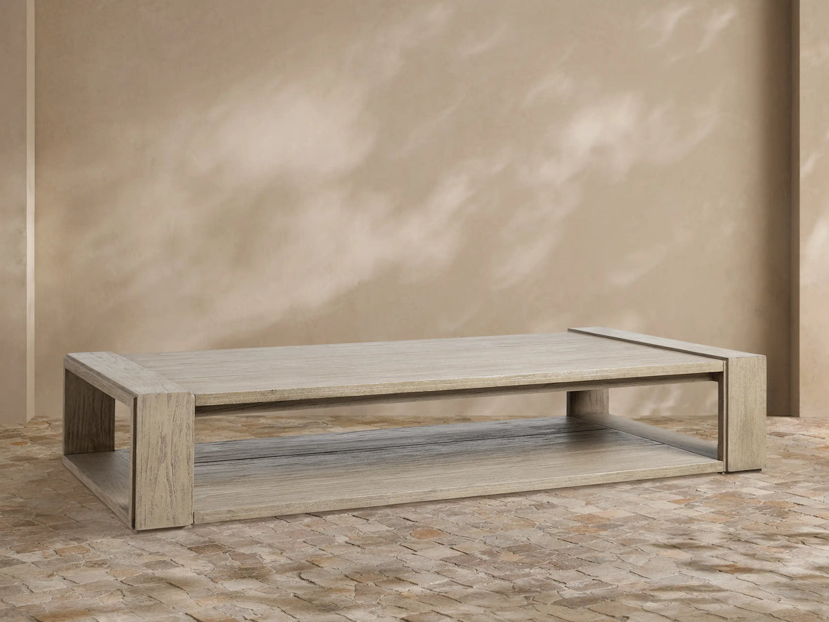 Canyon Outdoor Teak Coffee Table, rectangular, weathered grey teak, two-tiered, on a stone patio with beige wall.