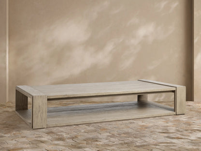Canyon Outdoor Teak Coffee Table, rectangular, weathered grey teak, two-tiered, on a stone patio with beige wall.