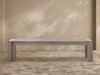 Canyon Outdoor Teak Dining Bench, weathered grey-brown wood, long rectangular seat & thick legs, on a patterned floor against a beige wall.
