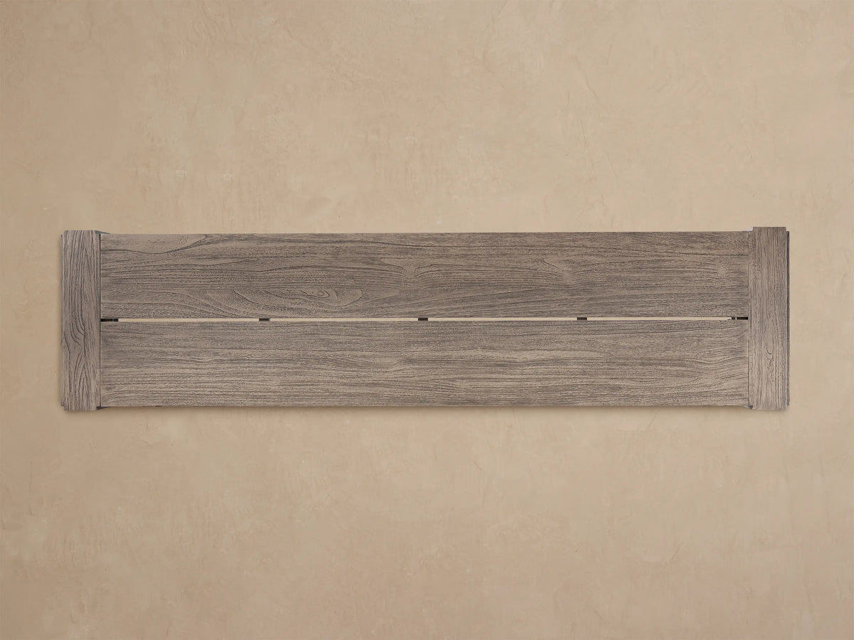 Canyon Outdoor Teak Dining Bench, weathered grey-brown wood, long rectangular seat & thick legs, on a patterned floor against a beige wall.