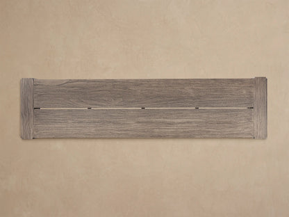 Canyon Outdoor Teak Dining Bench, weathered grey-brown wood, long rectangular seat & thick legs, on a patterned floor against a beige wall.