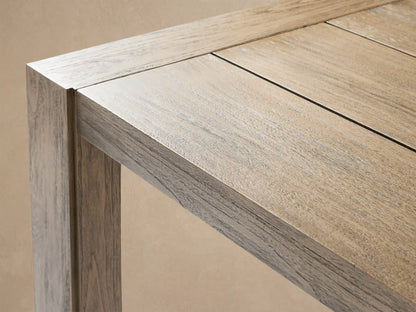 Canyon Outdoor Teak Dining Table, rectangular, light gray-brown distressed wood plank top, thick legs, against a beige wall on stone.