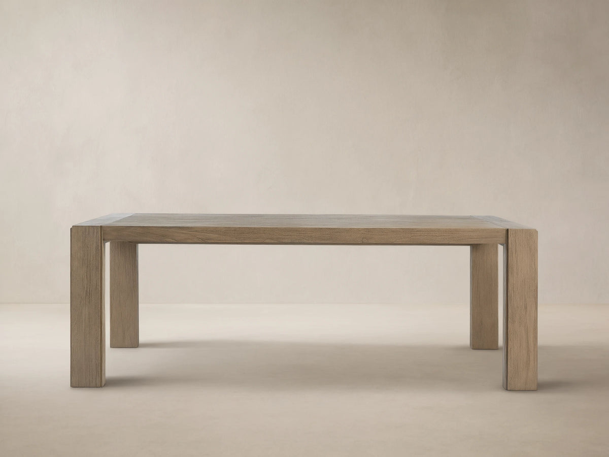 Canyon Outdoor Teak Dining Table, rectangular, light gray-brown distressed wood plank top, thick legs, against a beige wall on stone.