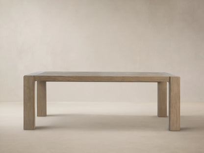Canyon Outdoor Teak Dining Table, rectangular, light gray-brown distressed wood plank top, thick legs, against a beige wall on stone.