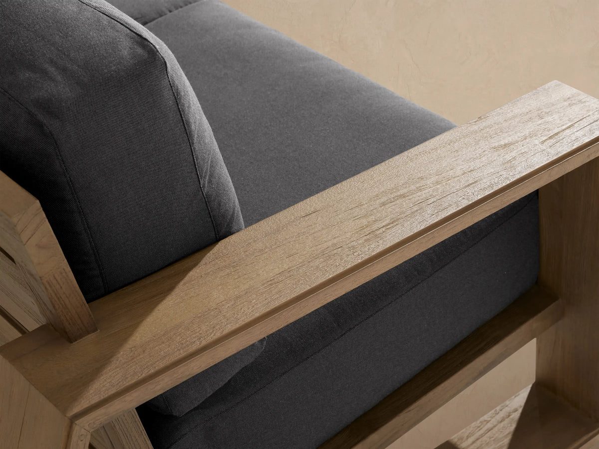 Canyon Outdoor Teak Sofa, angled, showing its natural teak wood frame and dark charcoal cushions in an outdoor patio.