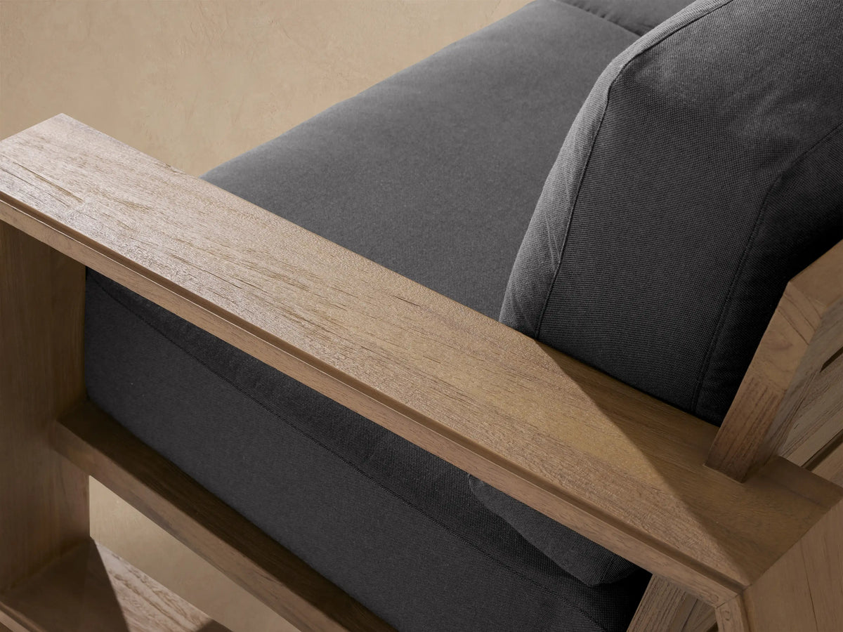 Canyon Outdoor Teak Sofa with natural wood frame & dark gray cushions, angled on a light tiled patio.