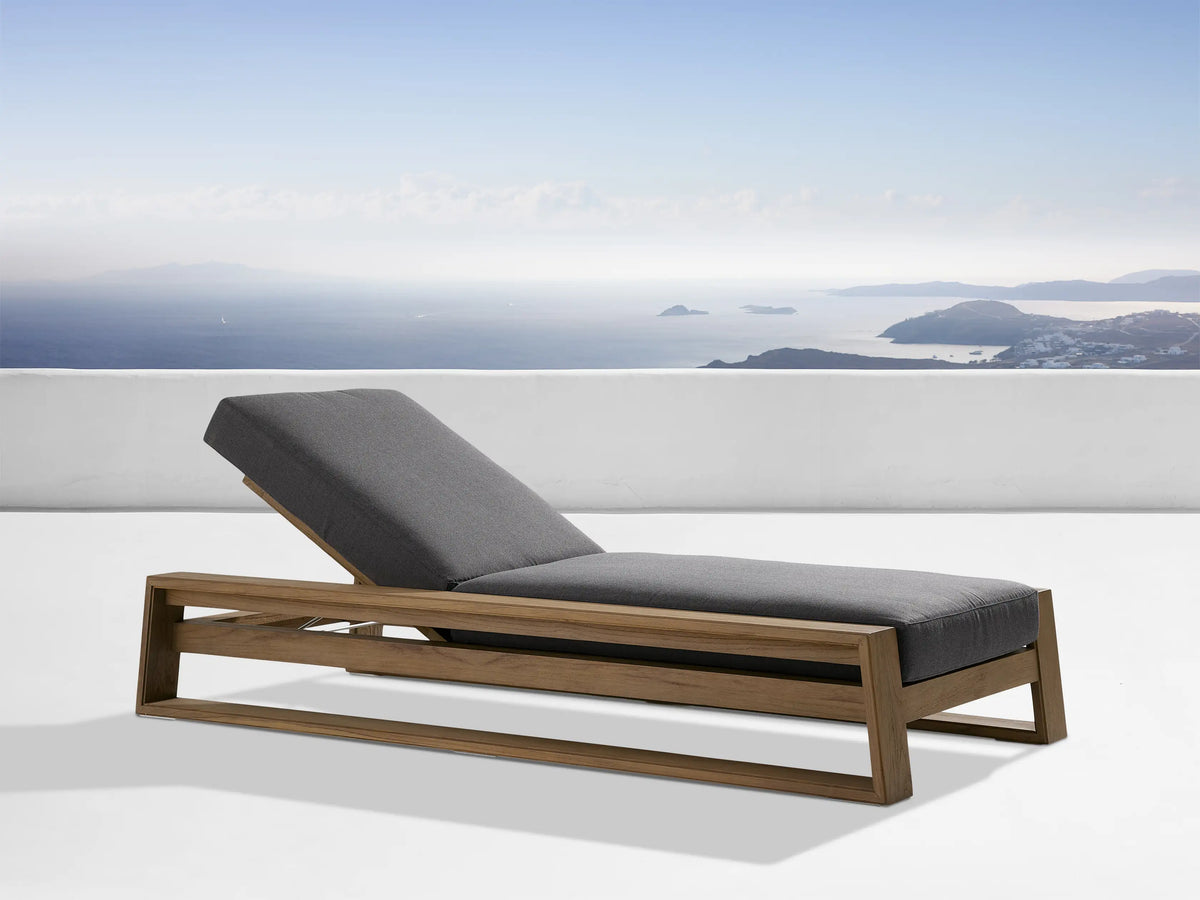 Canyon Outdoor Teak Chaise with an adjustable backrest in a natural teak finish. Gray cushions on a minimalist frame. Scenic ocean view backdrop.