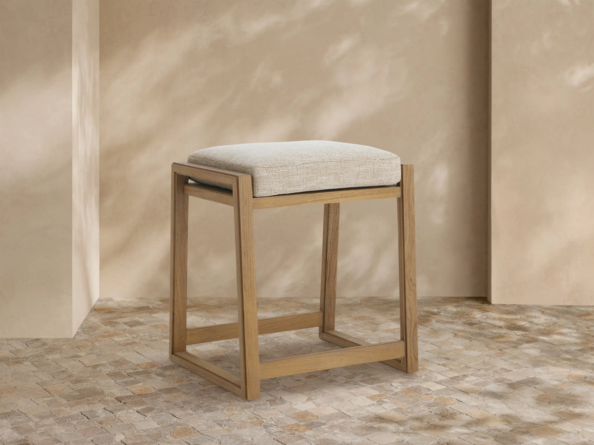Canyon Outdoor Teak Backless Counter Stool, light teak wood frame, soft Priano Malt cushioned seat, angled left on tiled floor.