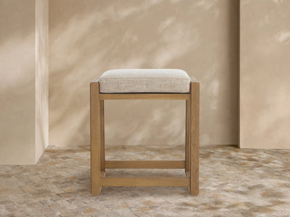 Canyon Outdoor Teak Backless Counter Stool, light teak wood frame, soft Priano Malt cushioned seat, angled left on tiled floor.