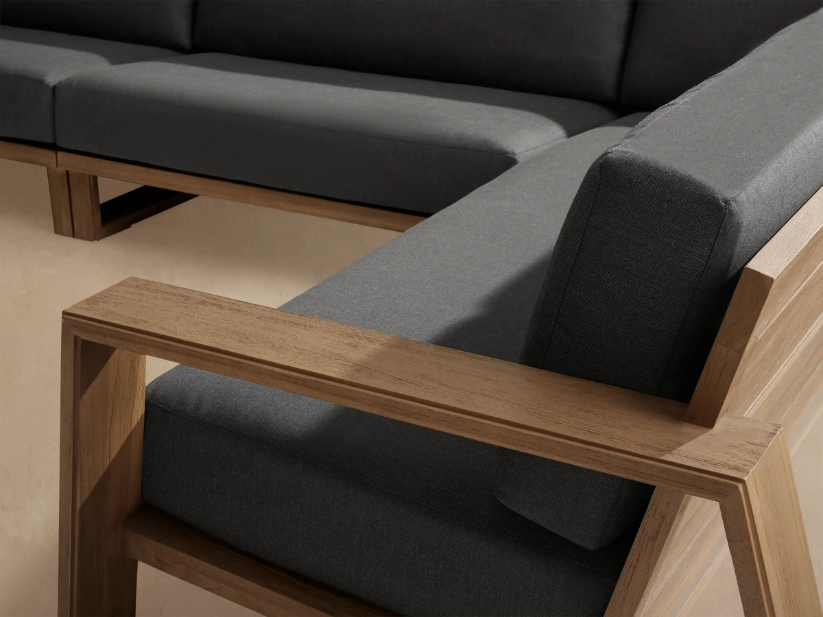 Canyon Outdoor Teak Four-Piece Sectional, L-shaped, with natural teak wood frame & dark grey cushions on a stone patio.
