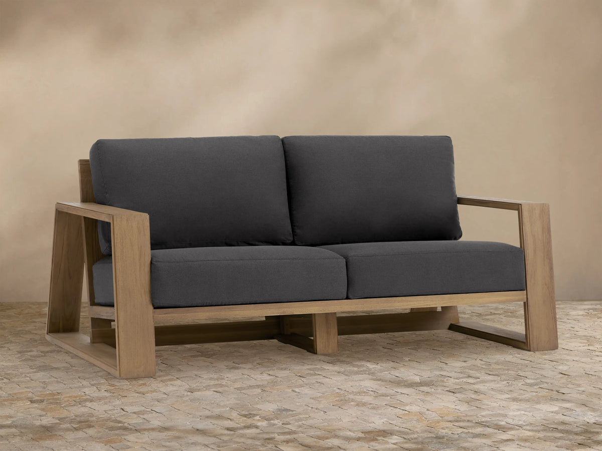 Front view of Canyon Outdoor Teak Sofa featuring a natural teak wood frame and dark gray cushions, set on a light-colored tiled patio.