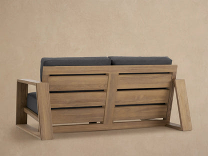 Angled close-up of Canyon Outdoor Teak Sofa: light brown, textured teak armrest & dark grey seat/back cushions.