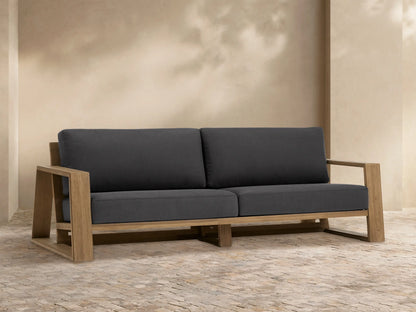 Front view of Canyon Outdoor Teak Sofa with natural teak wood frame and dark gray cushions, on a tiled patio.