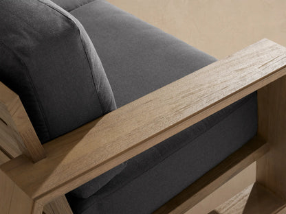 Canyon Outdoor Teak Sofa with light brown wood frame & dark gray cushions on a tiled patio against a tan wall.
