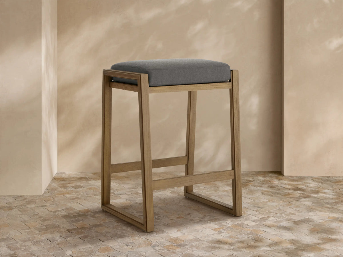 Canyon Outdoor Teak Backless Barstool: modern light teak wood frame, gray cushion, on a stone tile floor by a textured wall.