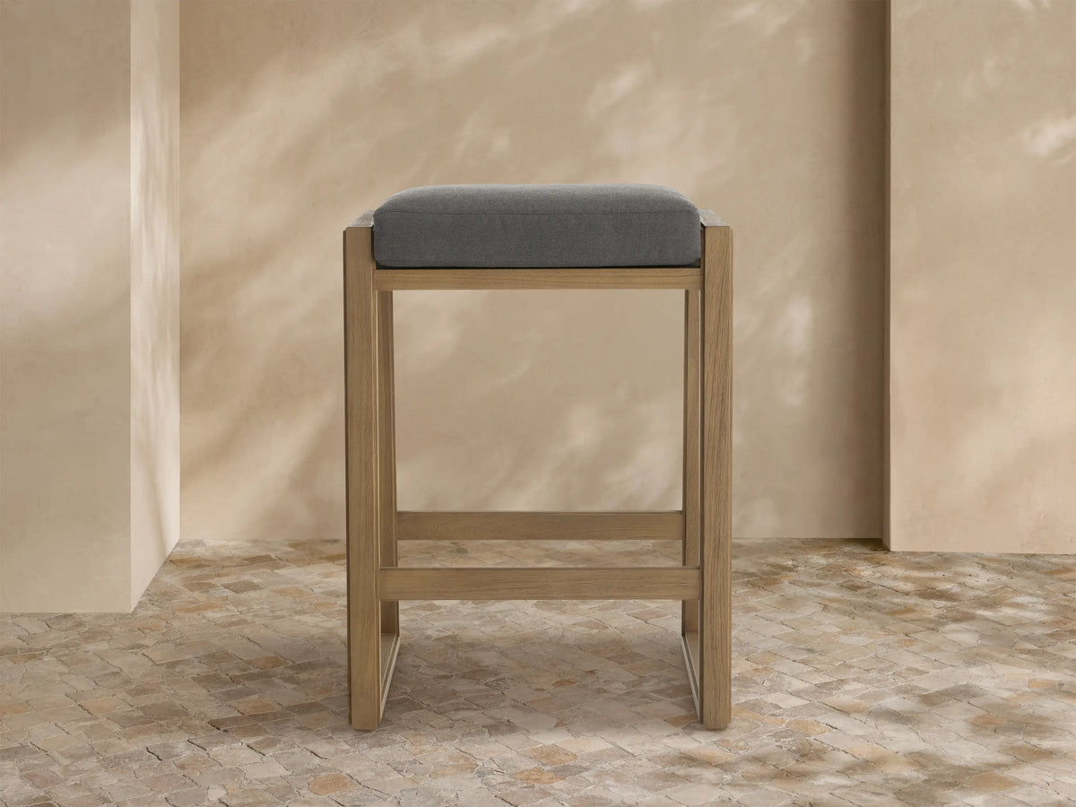 Canyon Outdoor Teak Backless Barstool: modern light teak wood frame, gray cushion, on a stone tile floor by a textured wall.