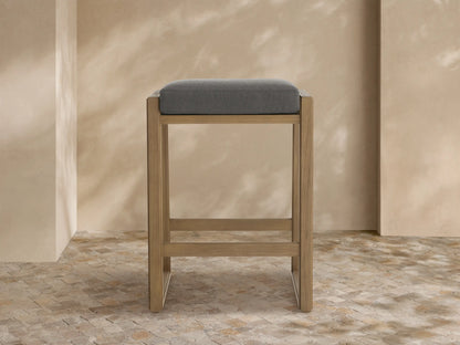 Canyon Outdoor Teak Backless Barstool: modern light teak wood frame, gray cushion, on a stone tile floor by a textured wall.