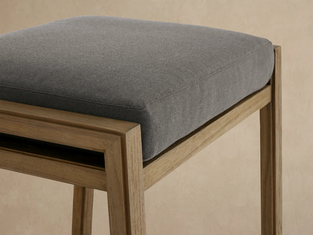 Canyon Outdoor Teak Backless Barstool with a light natural teak frame and dark grey cushion, against a beige wall & tiled floor.